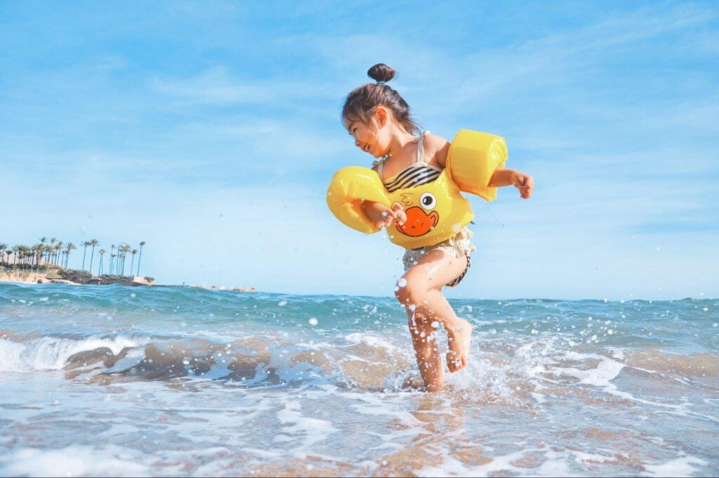 imagem com criança na praia, usando boias amarelas, brincando na água