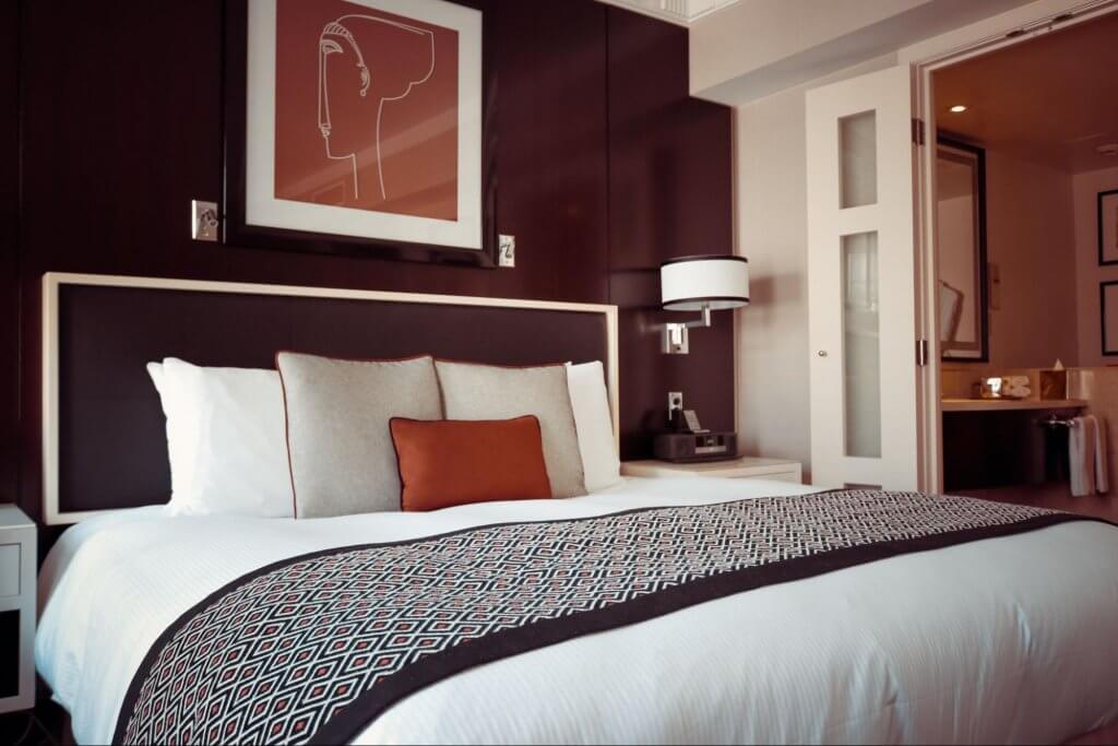 quarto de casal em hotel. Imagem mostra uma cama de casal, com almofadas e cobertas e um banheiro no segundo plano da imagem
