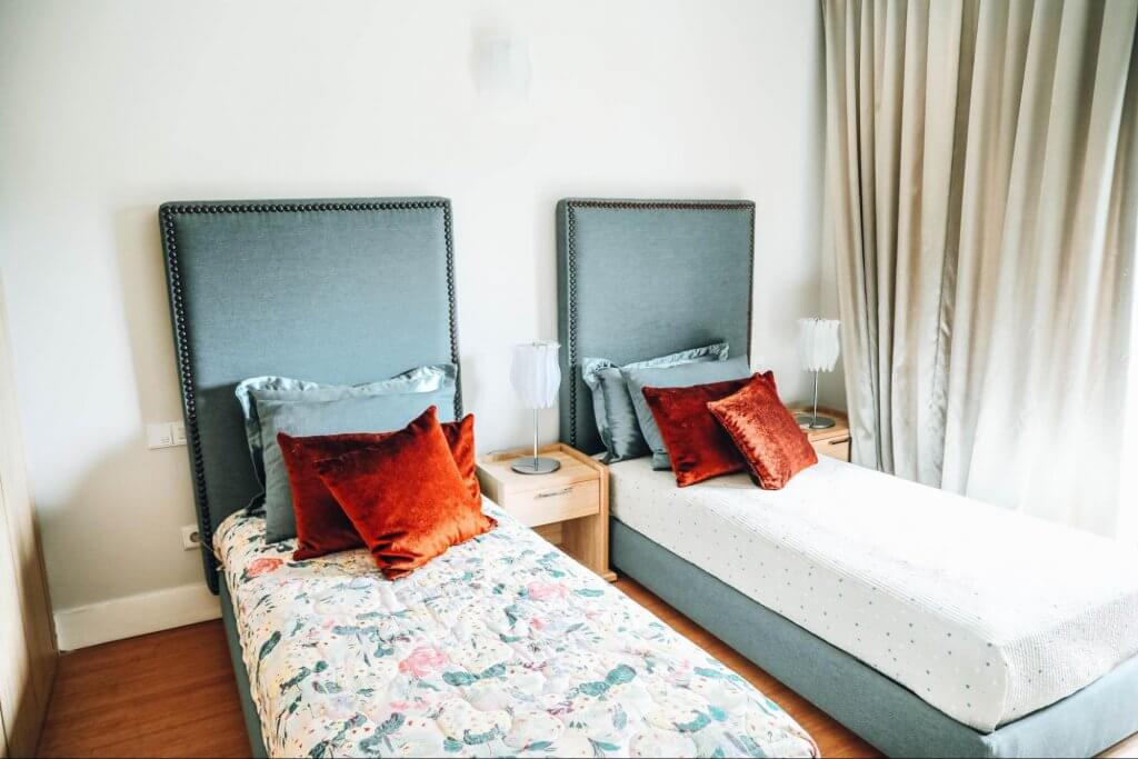 quarto de hotel duplo solteiro. A imagem mostra duas camas de hotel, de solteiro, separadas por uma mesinha de cabeceira. Por cima das camas, almofadas coloridas