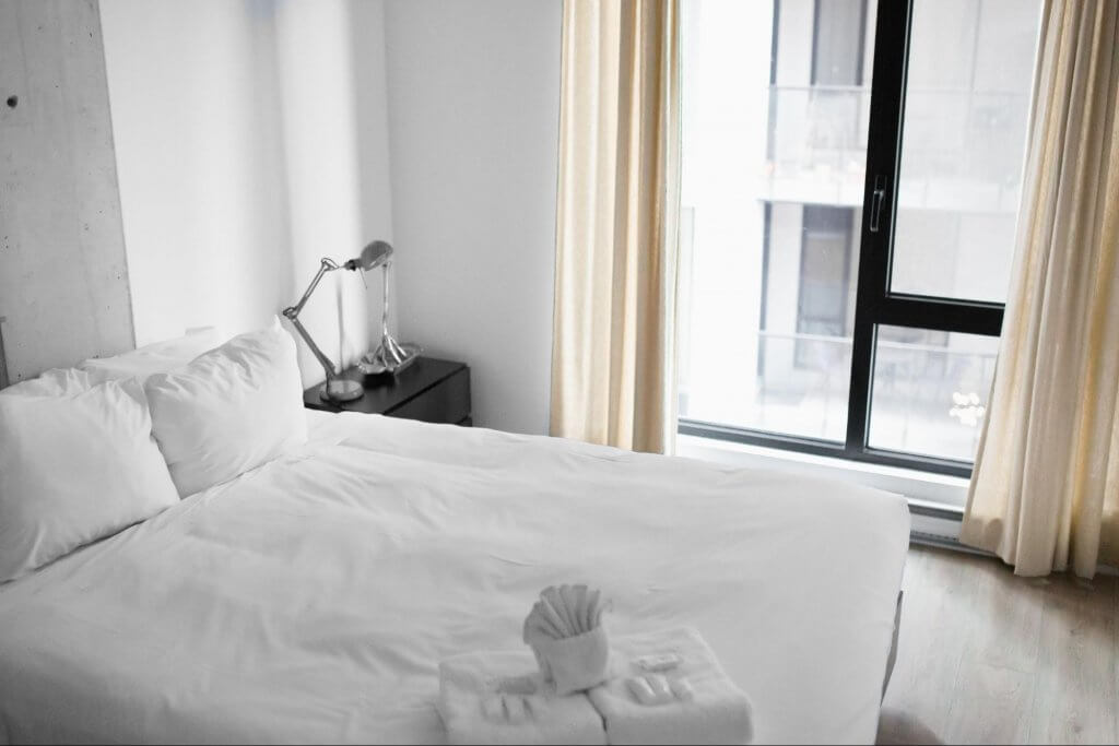 quarto de hotel solteiro. Imagem mostra uma cama, com travesseiro e toalhas de banho brancas. Ao fundo, janela com vista para cidade