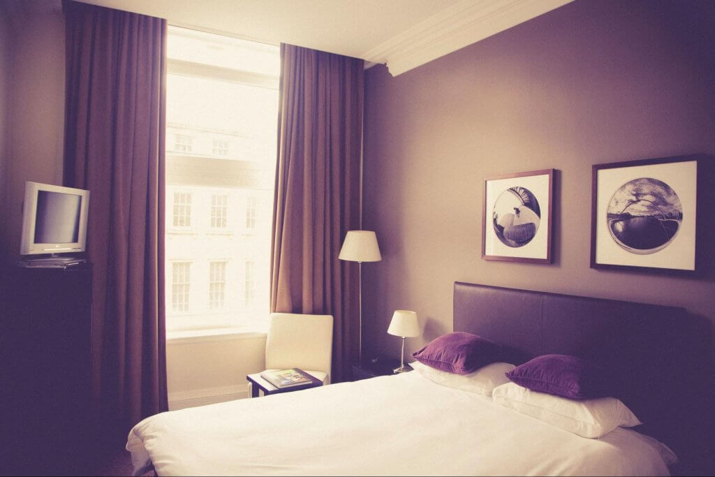 quarto de hotel standart. Imagem mostra cama de casal com travesseiros, mesinha lateral com abajur e uma pequena televisão presa na parede