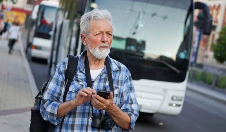 Idoso de camisa xadrez, andando na rua, com uma câmera pendurada no pescoço e o celular na mão. Atrás dele há um ônibus.