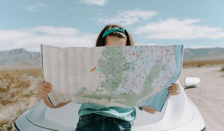 Mulher olhando um mapa de papel enorme e encostada no capô do carro