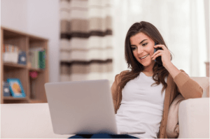 mulher usando casaco marrom por cima de camiseta branca sentada em um sofá comum laptop no colo enquanto fala ao telefone