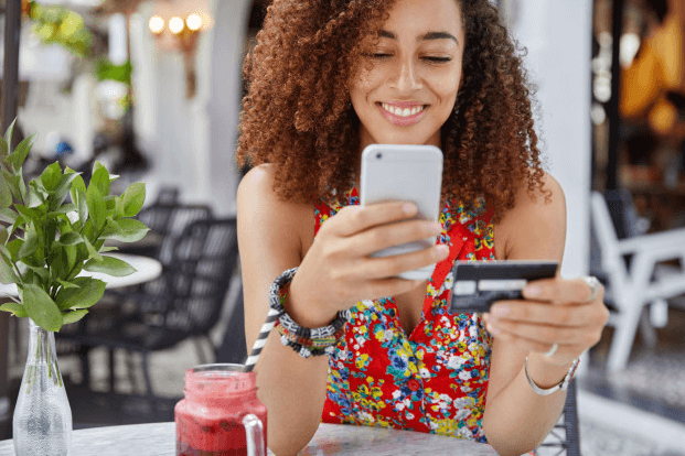 mulher sorrindo sentada à mesa mexendo no celular enquanto segura cartão de crédito