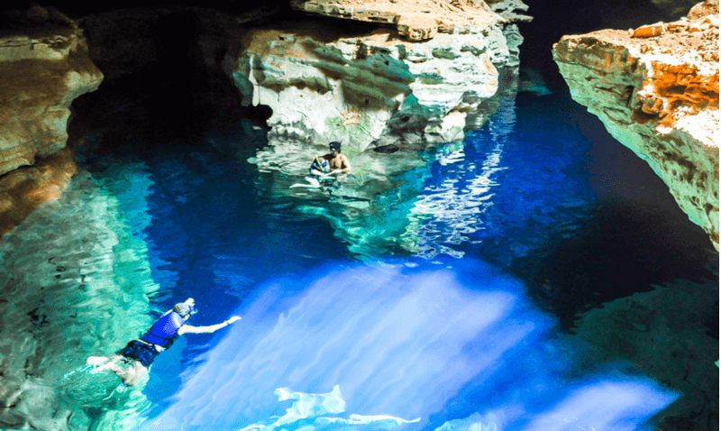 Imagem superior da Gruta Azul, com uma água de coloração forte na parte de baixo e muitas formações rochosas em volta. Duas pessoas fazem mergulho com snorkel.