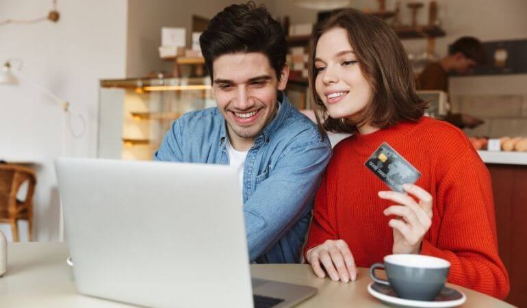 homem e mulher sentados em frente a um notebook. Ela segura um cartão de crédito e os dois estão sorrindo.